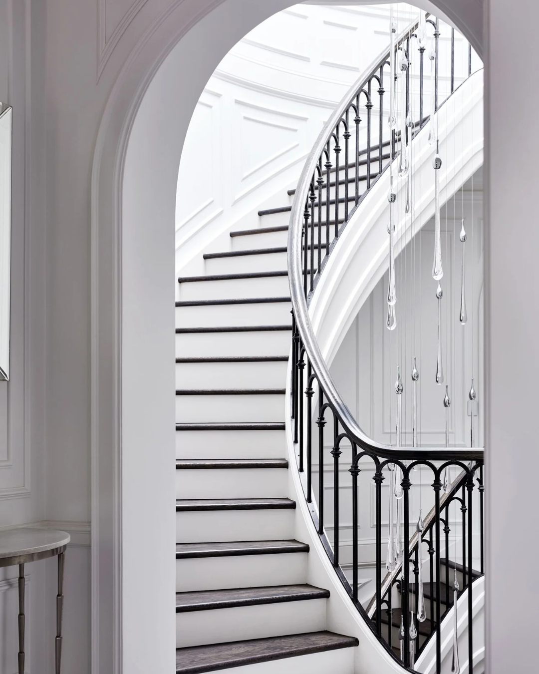 Bijou Custom Void Pendant Light cascading beside a sweeping curved staircase with a black wrought-iron balustrade in a luxury home