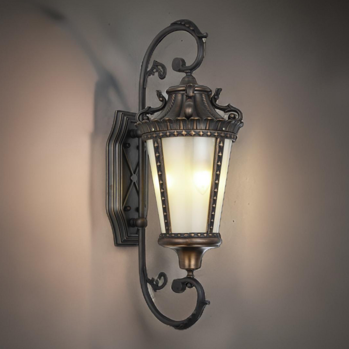 Bordeaux brass outdoor wall lamp with ornate French scrollwork and a sculpted glass lantern casting a warm glow on an exterior wall.