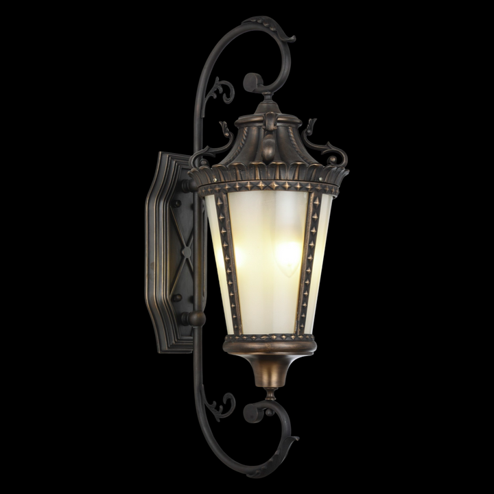 Bordeaux brass outdoor wall sconce featuring intricate French scrollwork and a sculpted glass shade in a heritage lantern design.