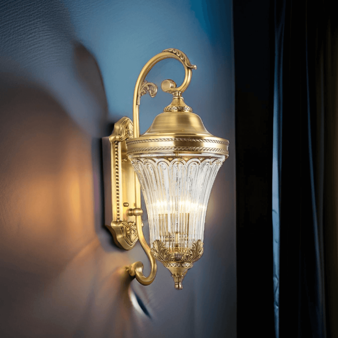 Rococo brass outdoor wall lamp with ornate European styling and a sculpted glass lantern casting a warm glow on an exterior wall.